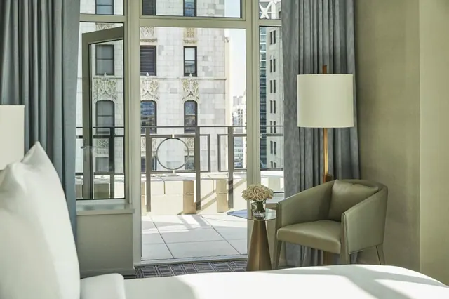 A bedroom with a made bed, a green armchair beside a floor lamp, and open glass doors onto a balcony overlooking an ornate stone building.