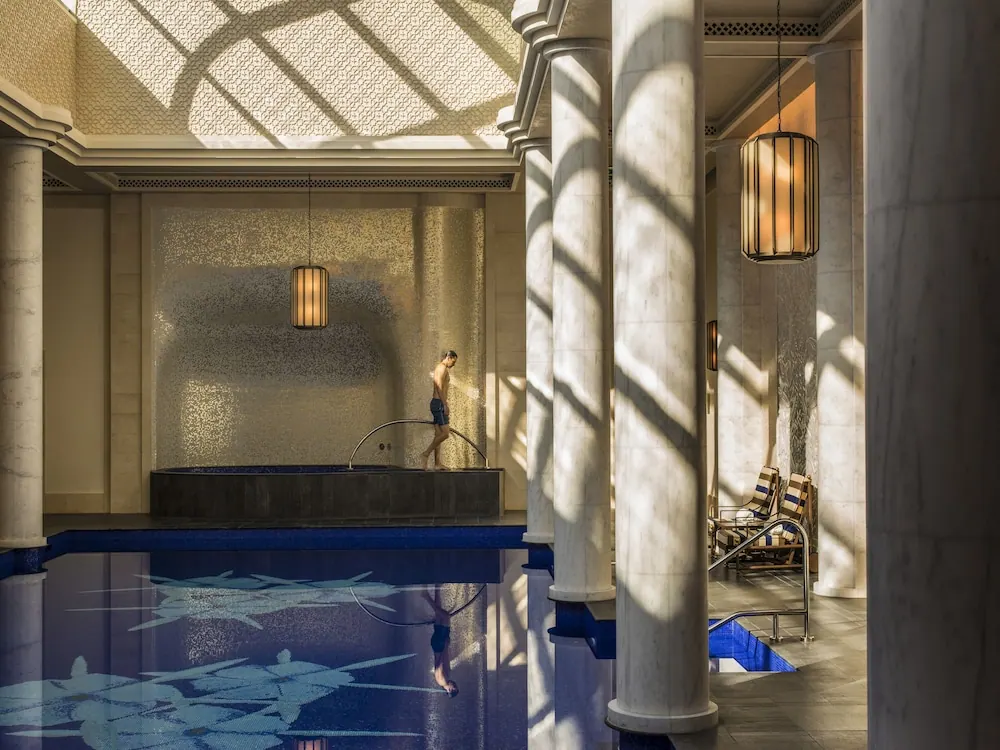 An indoor pool with blue mosaic tiles, marble columns and striped lounge chairs, with a man standing beside a sunken basin with a metal handrail.