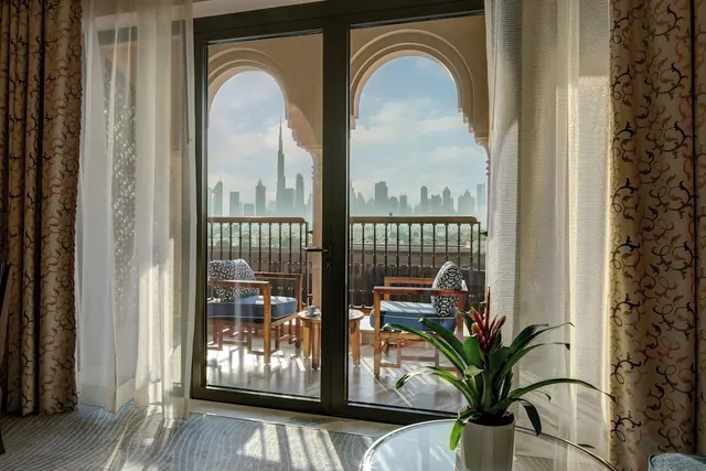 A balcony with wooden chairs and a small table is seen through glass doors framed by curtains, with the Burj Khalifa and city skyline in the background.