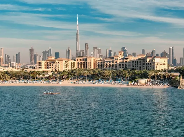 Seaward view of a beachfront hotel with sun loungers, palm trees and sandy beach, with the Burj Khalifa rising above the city skyline behind it.