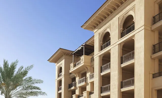 Exterior of a cream-coloured hotel building with arched balconies and iron railings, with a palm tree at left under a clear blue sky.