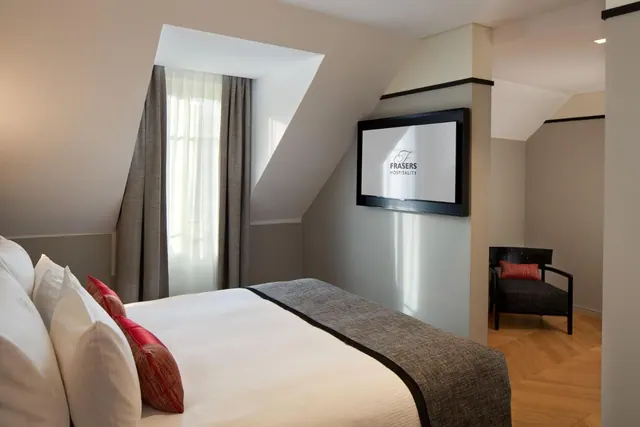 A bright attic-style hotel bedroom has a neatly made double bed, television, tall curtained window and armchair.