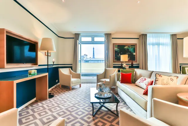 A furnished sitting room has a television, sofas, armchairs, and a clear view of the Eiffel Tower through the window.