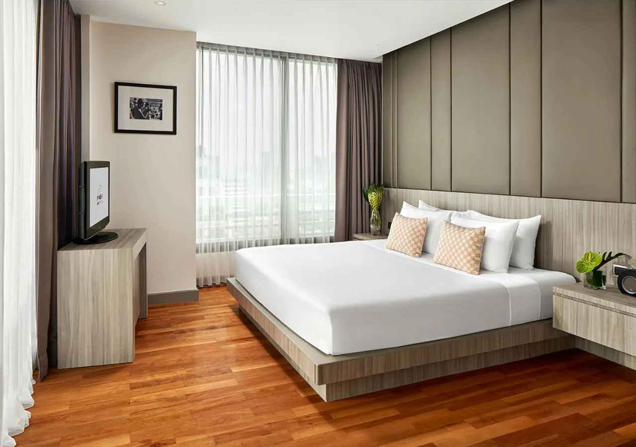 A bedroom with a large white bed, a television on a wooden unit, and a window with sheer curtains looking out over a hazy cityscape.