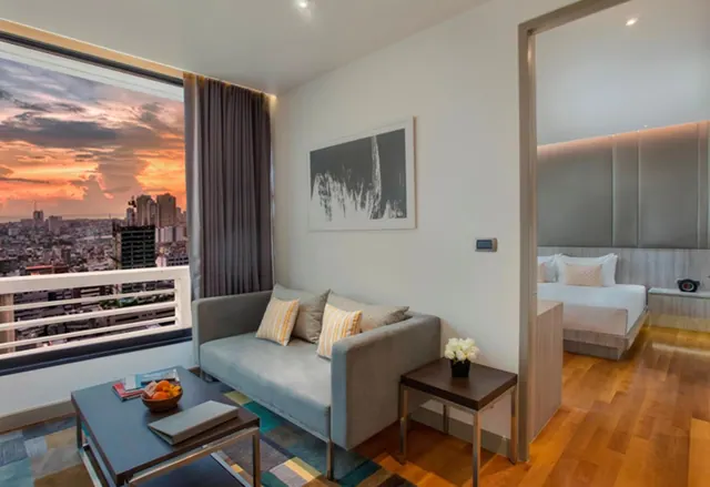 A living area with a grey sofa, coffee table and large window showing a city skyline at sunset, with a bedroom visible through the doorway.