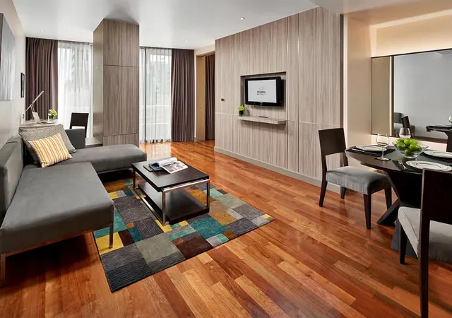 A living room with a grey sofa, a dark coffee table on a patterned rug, a wall-mounted TV, and a round dining table with chairs beside floor-to-ceiling windows.