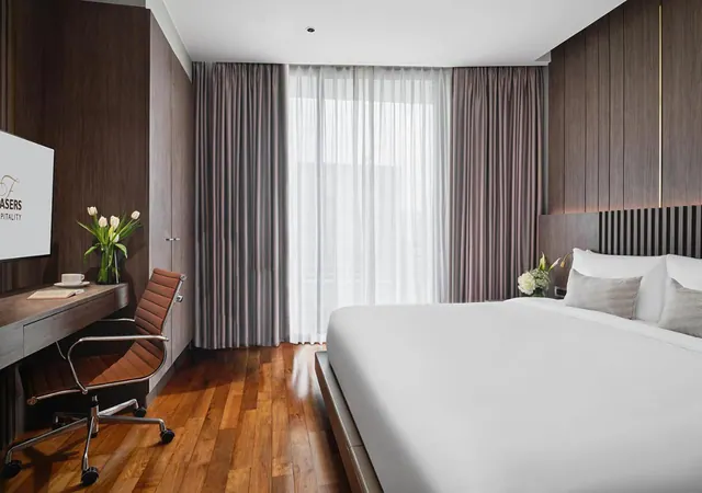 A made-up king bed with white linens sits across from a wooden desk, leather chair and wall-mounted television, with sheer curtains over a window.