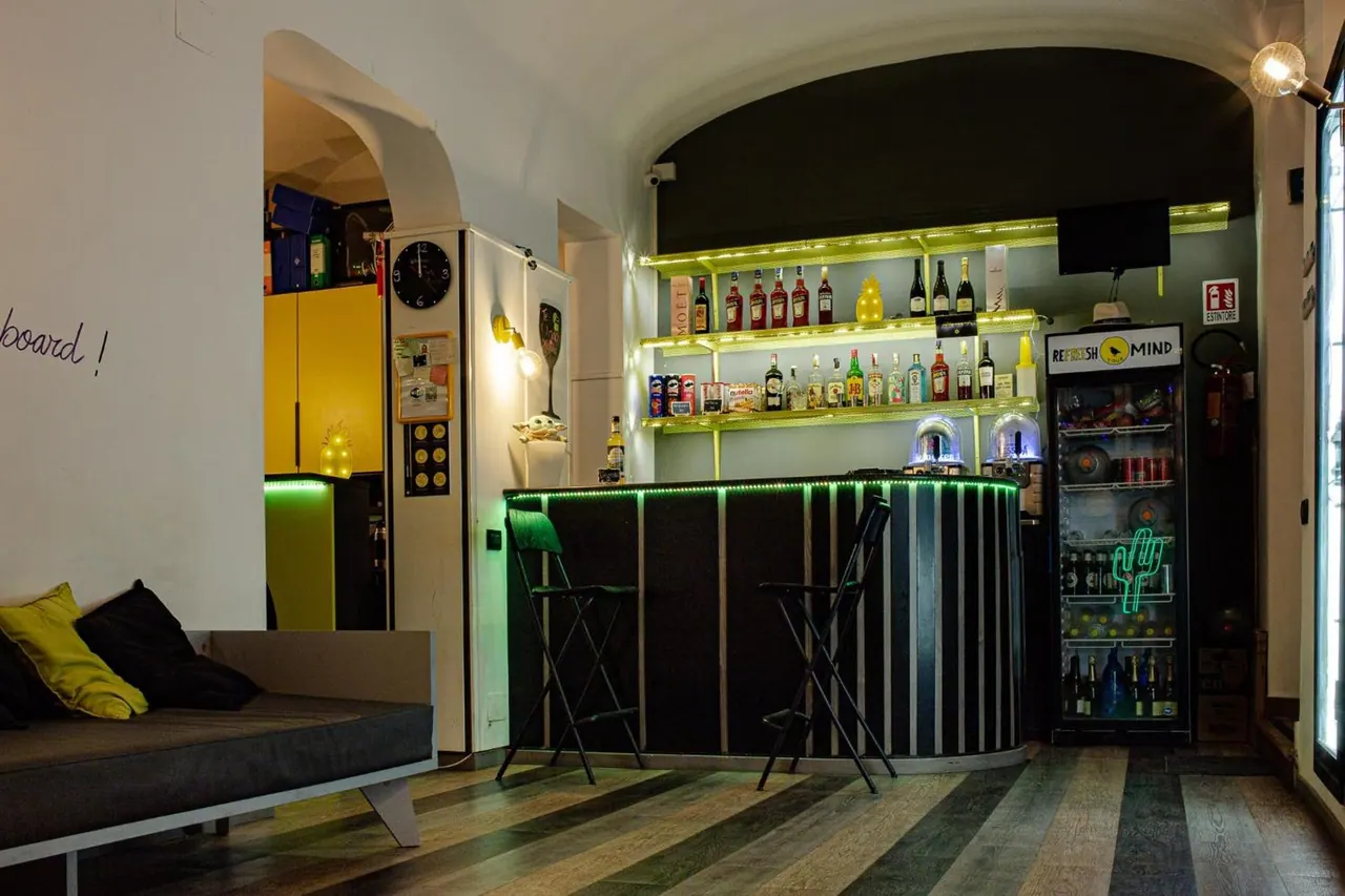 A bar counter with green LED lighting, illuminated bottle shelves, two high stools, a drinks fridge and a grey sofa with cushions in a lounge area.