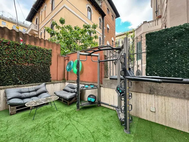 An outdoor courtyard with artificial turf, a grey cushioned sofa and armchair on wooden pallets, a small glass-topped table, a metal workout rack with medicine balls, and hedges along the walls.