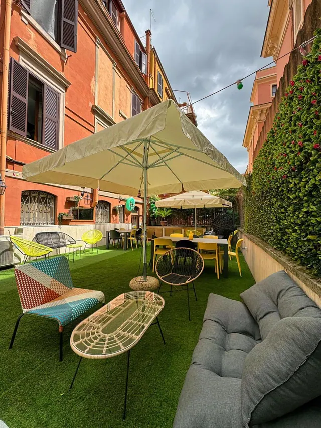 An outdoor terrace with artificial grass, shade umbrellas, colourful chairs, a grey cushioned sofa and a tall green hedge wall beside an orange building.