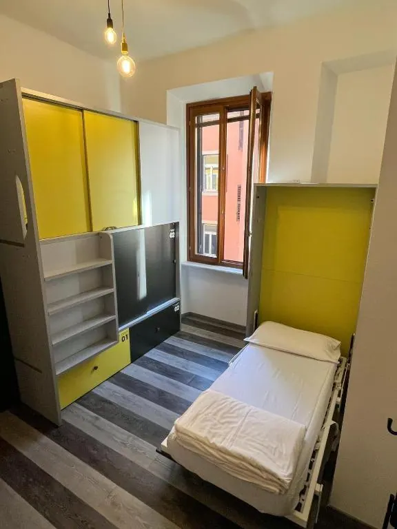 A small bedroom with a pulled-out fold-down bed, yellow wardrobe doors, open shelving, a striped wood-effect floor, and a window looking onto a building across the street.