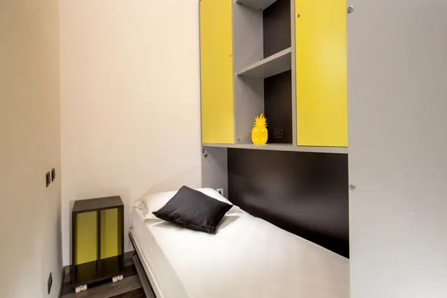 A small bedroom with a single bed, a black pillow, yellow wall cabinets and a yellow pineapple ornament on a shelf.