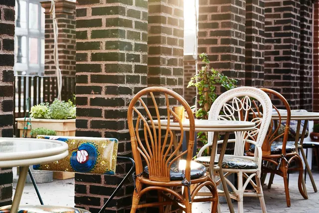 An empty outdoor seating area with rattan chairs, round tables and planters beside tall brick pillars, lit by low sun.