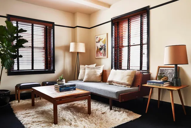 A living area with a grey upholstered sofa, a wooden coffee table with books on it, a shag rug, two table and floor lamps, and windows with wooden blinds.