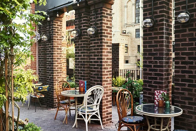 An outdoor terrace with brick pillars, round glass-topped tables, wicker chairs with blue cushions and hanging string lights.