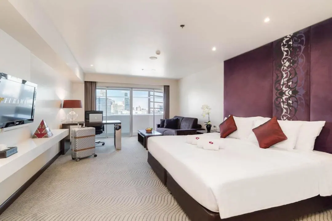 A hotel bedroom with a large white bed against a purple headboard wall, a sofa, a desk, a wall-mounted television and sliding glass doors showing city towers.