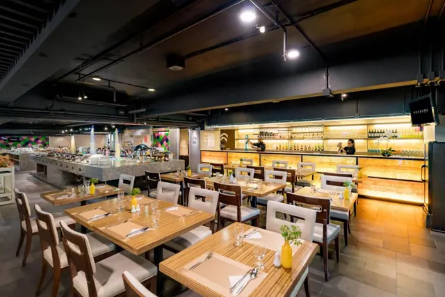 A restaurant dining room with wooden tables and white chairs, a backlit bar with bottles on the right, and a buffet station along the left wall.