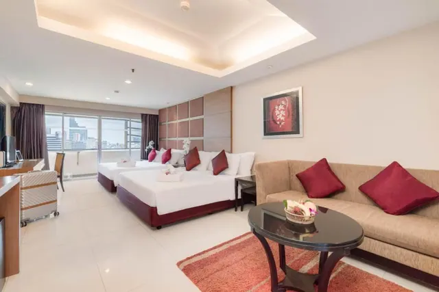 A hotel room with two beds with red cushions, a sofa and round coffee table, and a window showing city buildings.