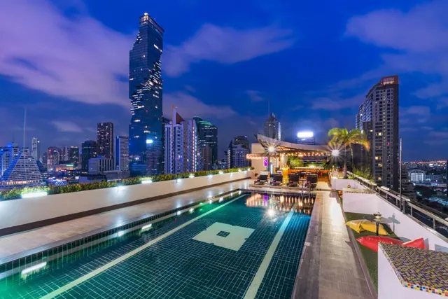 A rooftop pool at dusk with a bar and sun loungers at the far end, with the MahaNakhon tower and city skyline behind.