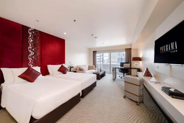 A hotel room with two single beds under a red padded headboard, a sofa, a desk with a chair, a wall-mounted television and a suitcase near the window showing city buildings.