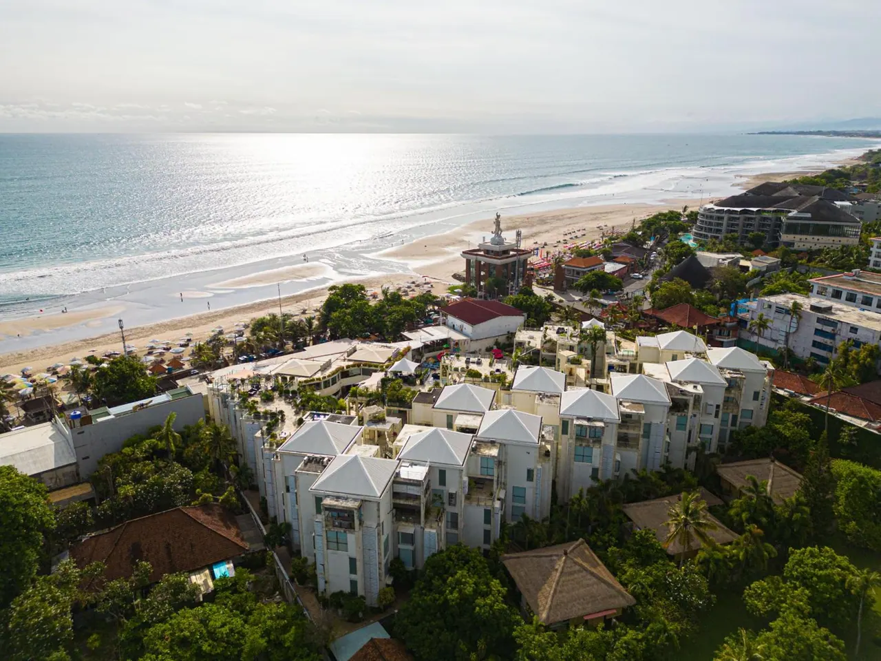 Aerial view of a white multi-storey hotel complex with grey roofs beside a sandy beach and the ocean.