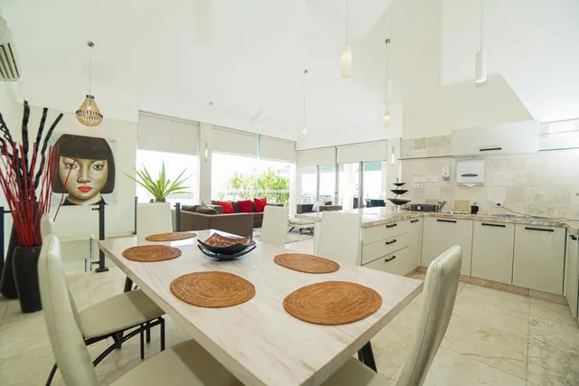 A bright open-plan living and dining area with a wooden dining table, white chairs, a modern white kitchen and a sofa with red cushions beyond, with greenery visible through the windows.