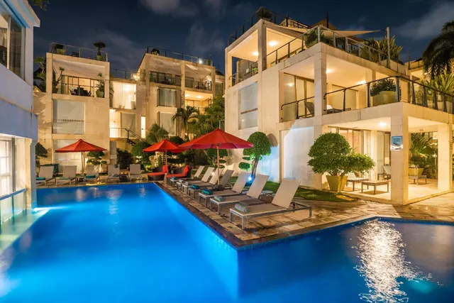 An illuminated swimming pool at night with red umbrellas and sun loungers, beside multi-storey buildings with balconies.