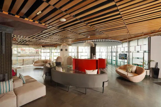 A hotel lobby lounge with curved grey sofas, a red reception counter, hanging rattan chairs, chandeliers and a wooden slat ceiling.