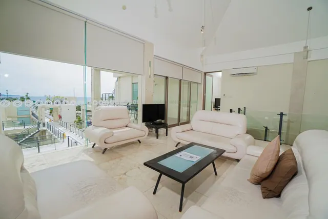 A bright living area with white leather sofas, an armchair, a glass-topped coffee table, a television and a view through large windows toward the sea.