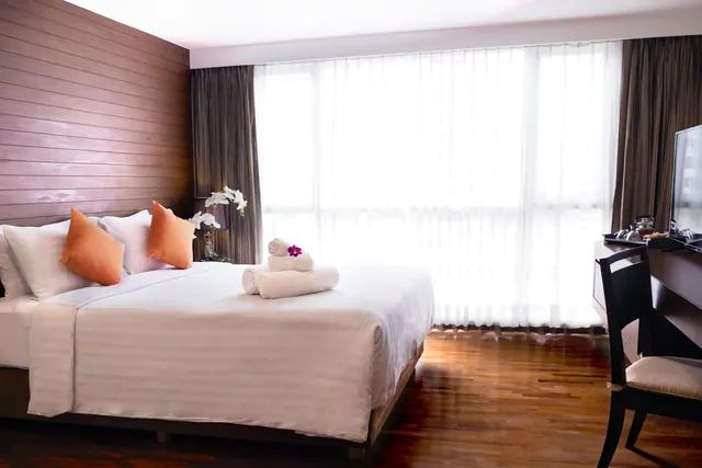A bedroom with a made-up double bed, folded towels with an orchid, a wooden desk with a television, and sheer curtains over a large window.