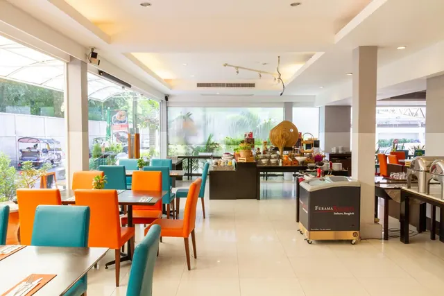A bright dining room with orange and teal chairs and set tables, a buffet counter in the background, and large windows onto a street where a tuk-tuk is parked.