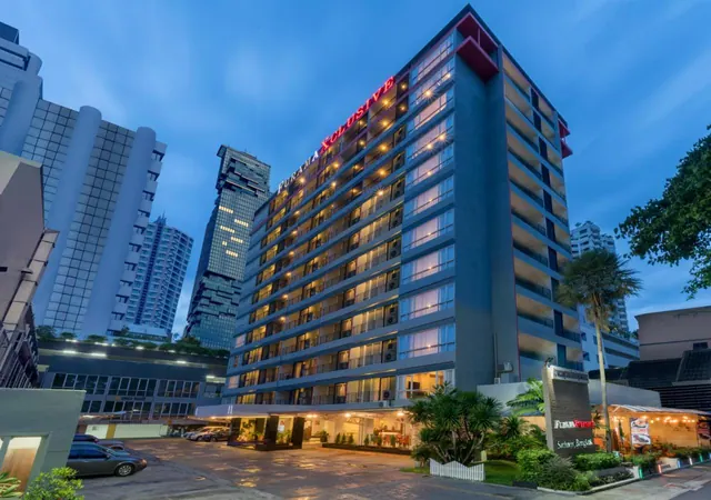 Exterior of a tall hotel tower at dusk with a red illuminated sign on its facade, a palm tree, and a parked car in the forecourt.