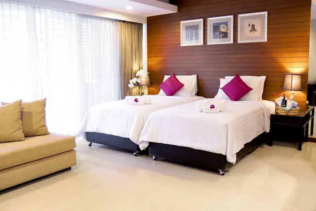 A bedroom with two beds dressed in white linen and magenta cushions, a beige sofa on the left, a wood-panelled wall with three framed prints, and sheer curtains over a large window.