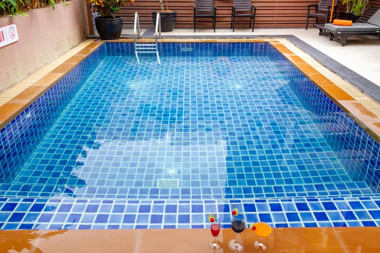 A small rectangular pool with blue mosaic tiles and a metal ladder, with three cocktail glasses on the edge in the foreground and sun loungers beyond the far side.