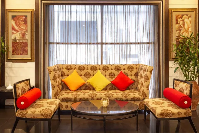 A lobby seating area with a patterned sofa, two armchairs with red bolster cushions, a round coffee table and framed artwork on the walls.