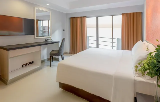 A bedroom with a made king bed, a wall-mounted television, a desk with a chair and mirror, and sliding doors onto a small balcony.