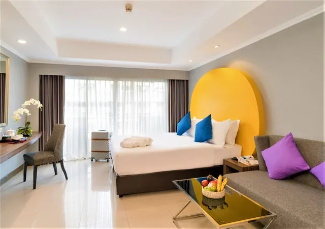A hotel bedroom with a king bed and a yellow arched headboard, a grey sofa with purple cushions, and a desk with a chair beside sheer-curtained windows.