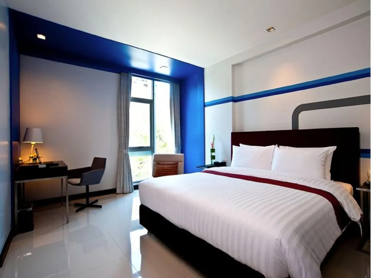A bedroom with a large bed with white striped linens, a desk with a lamp and chair, a blue accent wall, and a window with trees visible outside.