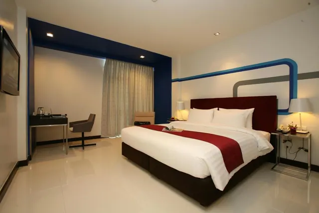 A bedroom with a large bed with a red runner, a desk with a chair and kettle, a grey armchair, and a wall-mounted television.