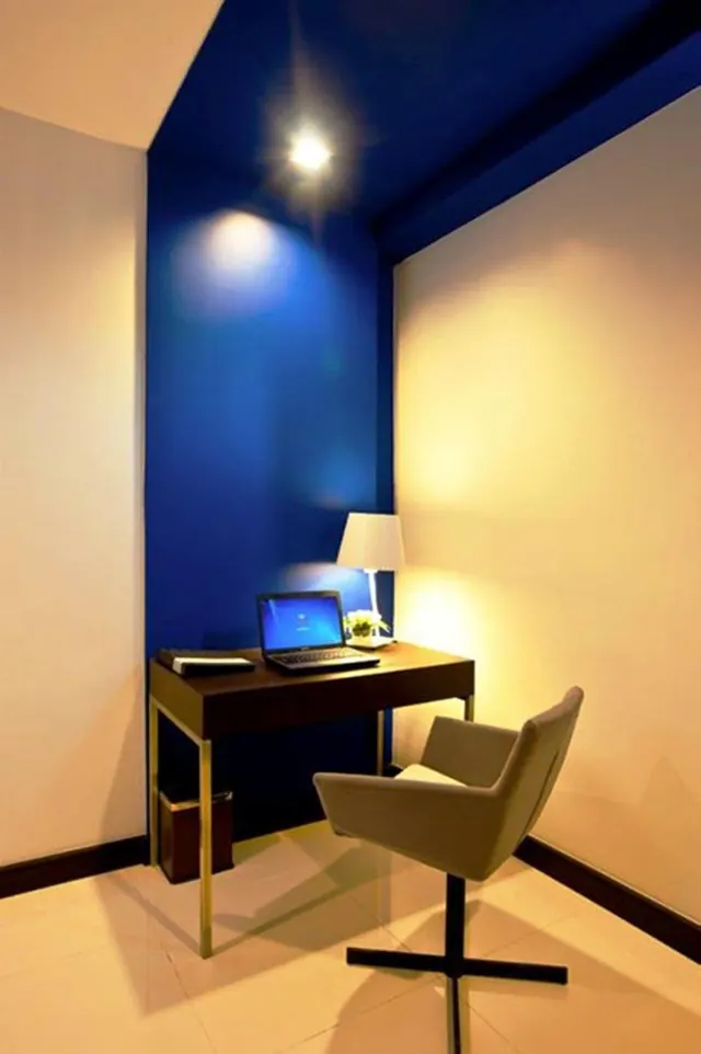 A corner workspace with a dark wooden desk, an open laptop, a table lamp and a grey swivel armchair against a deep blue wall.