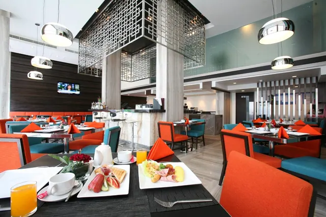 A restaurant dining room with orange and teal chairs, pendant lights, and breakfast plates set on a table in the foreground.