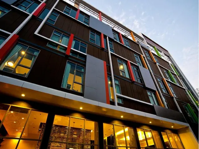 Low-angle dusk view of a multi-storey building facade with brown cladding, red, orange and green accent panels, and a lit ground-floor glass frontage.