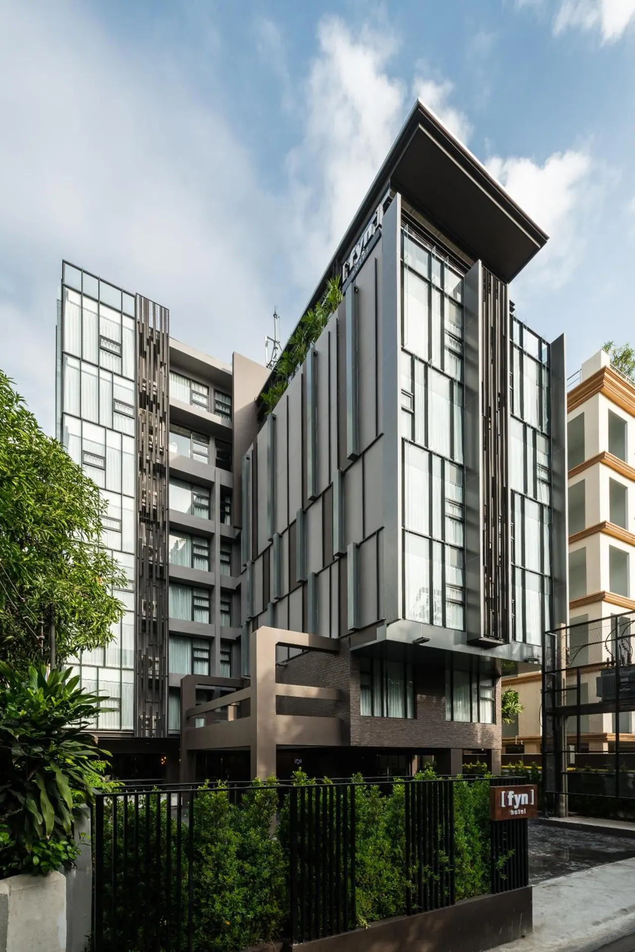 Exterior of the [fyn] hotel building, a multi-storey block with glass and dark metal cladding, with greenery and a black metal fence at street level.