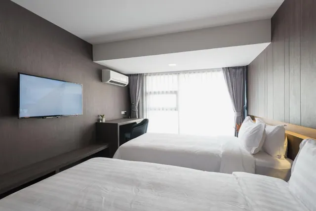 A hotel bedroom with two single beds, a wall-mounted television, a wall air conditioner and a desk with a chair beside a window with sheer curtains.