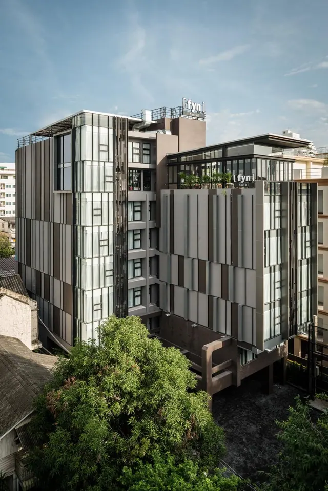 Exterior of the fyn hotel building, a multi-storey structure with glass and grey panelled facades, with a large tree in the foreground under a blue sky.