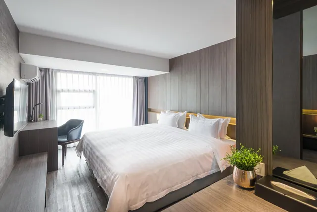 A bedroom with a king bed, a wall-mounted television, a desk with a chair, and a large window with sheer curtains.