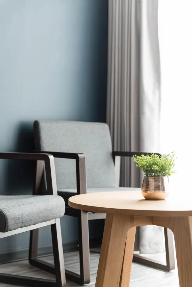 Two grey upholstered armchairs with dark wooden frames sit beside a round wooden side table holding a potted plant, with grey curtains and a blue wall behind.