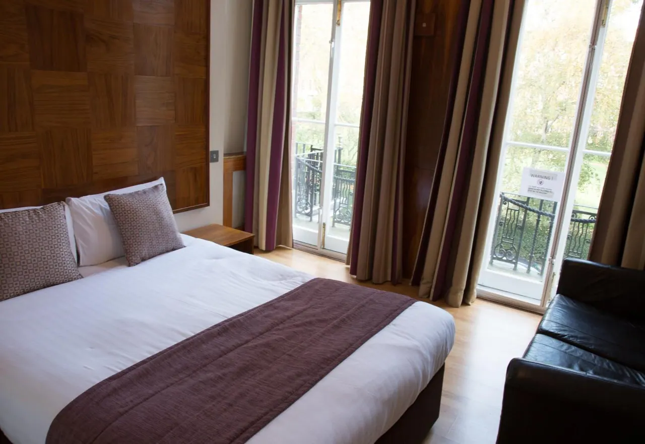 A double bed with a wooden panel headboard and purple throw sits beside French doors opening onto a balcony with trees beyond, next to a black leather sofa.
