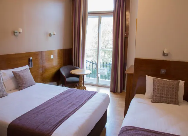 A hotel bedroom with a double bed and a single bed, an armchair and small round table beside a glazed door opening onto a small balcony.