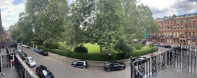 A panoramic view from a balcony over a tree-lined garden square and street with parked cars and red brick terraced buildings.